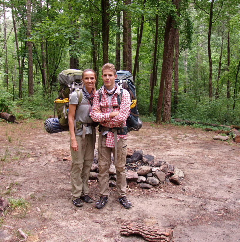 A woman and a young man stand side-by-side in a forest clearing, both wearing hiking attire and large backpacks. The woman is on the left, and the man is on the right with his arms crossed. Behind them, there's a circle of stones, suggesting a former campfire site. The area is surrounded by tall trees, creating a dense, green backdrop.